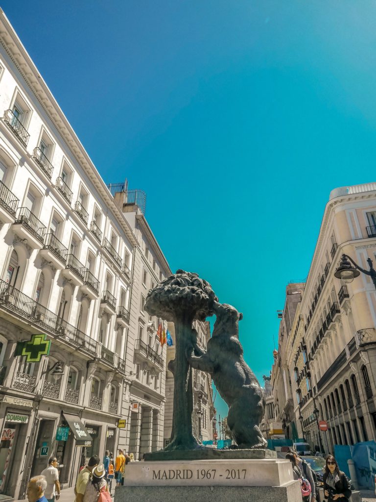 A statue of the symbol of Madrid (a bear reaching up to a hackberry tree), located in the middle of a city street with blue skies in the background